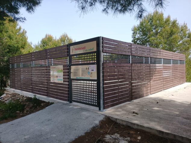 Imagen: Recinto cerrado de listones oscuros con puerta metálica y carteles informativos, rodeado de pinos.