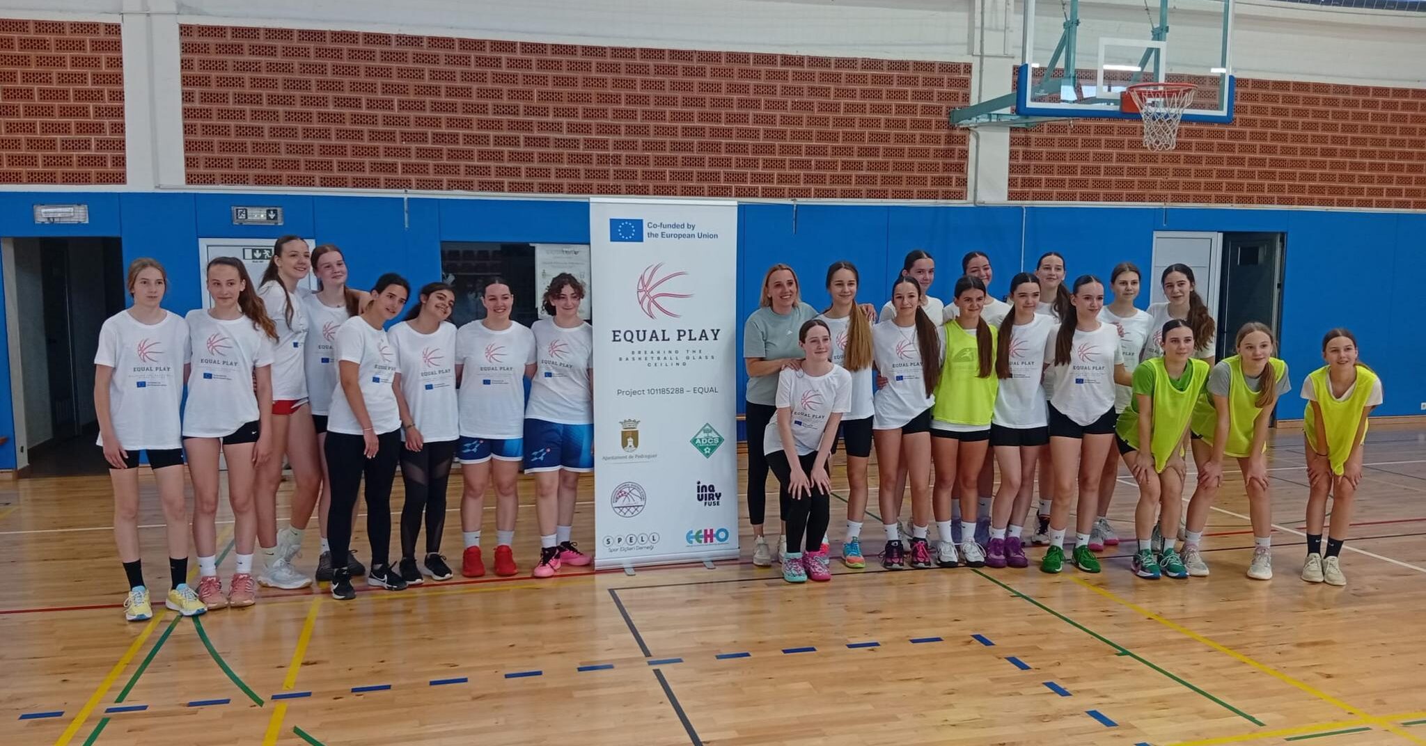 Foto de grupo de jóvenes jugadoras de baloncesto y entrenadores en un gimnasio, posando junto a un cartel del proyecto europeo EQUAL PLAY.