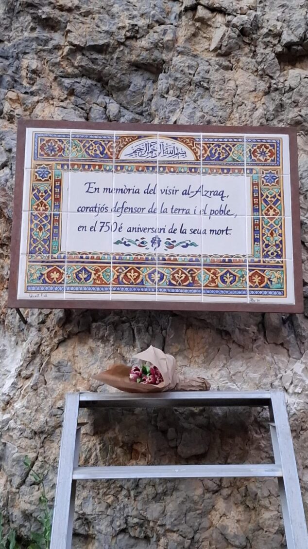 Imagen: Fotografia de la placa de ceràmica commemorativa del 750 aniversari de la mort del visir Al-Azraq, instal·lada en una paret de roca, amb un ram de flors depositat a sota sobre una escala metàl·lica.