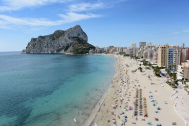 Vista aérea de la concurrida playa de la Fossa en Calp, con el majestuoso Peñón de Ifach dominando el paisaje costero bajo un cielo azul.