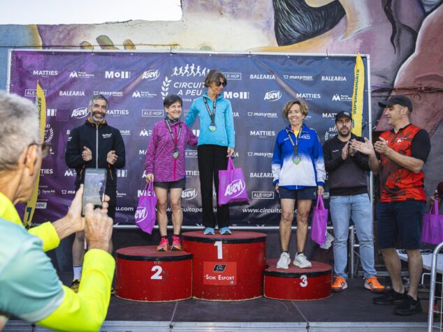 Imagen: Tres mujeres en el podio de la categoría Veteranas G femenina de la Cursa Jesús Pobre, sosteniendo bolsas de premios Asevi y recibiendo aplausos.