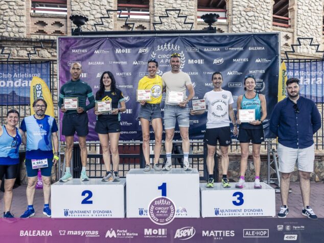Podio con tres corredores premiados sosteniendo trofeos y tres organizadores de pie a los lados, con un fondo de patrocinadores en la Cursa Popular Ondara.