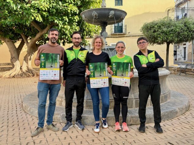 Imagen: Miembros del Club Atletisme Cametes y la concejala de Deportes posan con el cartel de la XXXIII Cursa Popular Vila de Pedreguer frente a una fuente.