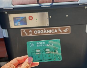 Benissa activa el reparto de tarjetas para sus nuevos contenedores inteligentes 3 Mano sosteniendo una tarjeta verde de acceso frente a un contenedor de basura orgánica gris con cerradura electrónica.