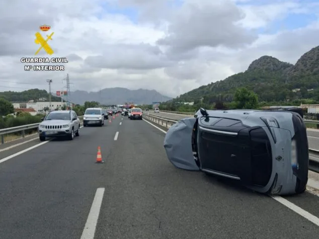 Imatge: Un cotxe gris bolcat al voral de l'autopista AP7, amb trànsit detingut al fons i el logo de la Guàrdia Civil.