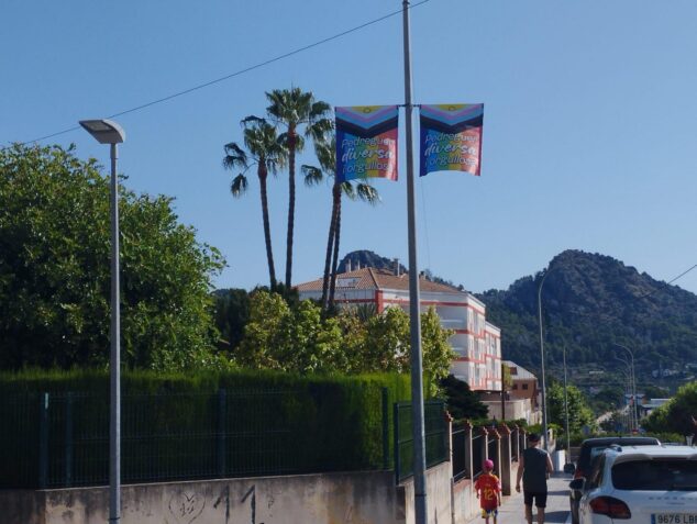 Imagen: Dos banderolas con los colores del arcoíris y el texto "Pedreguer diversa i orgullosa" colgadas en una farola en una calle.