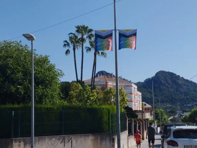 Imagen: Dos banderolas con los colores del arcoíris y el texto "Pedreguer diversa i orgullosa" colgadas en una farola en una calle.