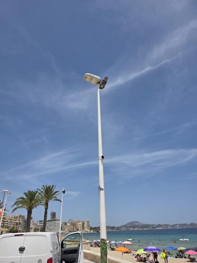 Imagen: Un poste con dispositivos de medición meteorológica en una playa de Calp, con el Peñón de Ifach y edificios al fondo bajo un cielo azul.