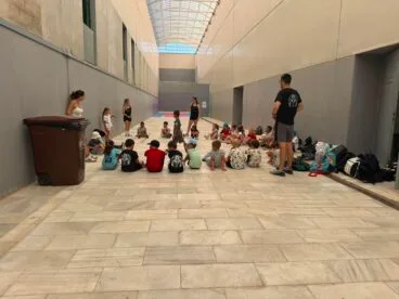 Grupo de niños sentados en el suelo atendiendo a monitores en un pasillo de instalaciones deportivas durante el campamento de verano de Calp.