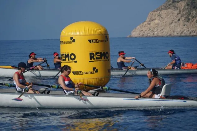 Imagen: Remeras compitiendo en el mar con una gran boya amarilla 'LIGA suma' y una colina rocosa de fondo.