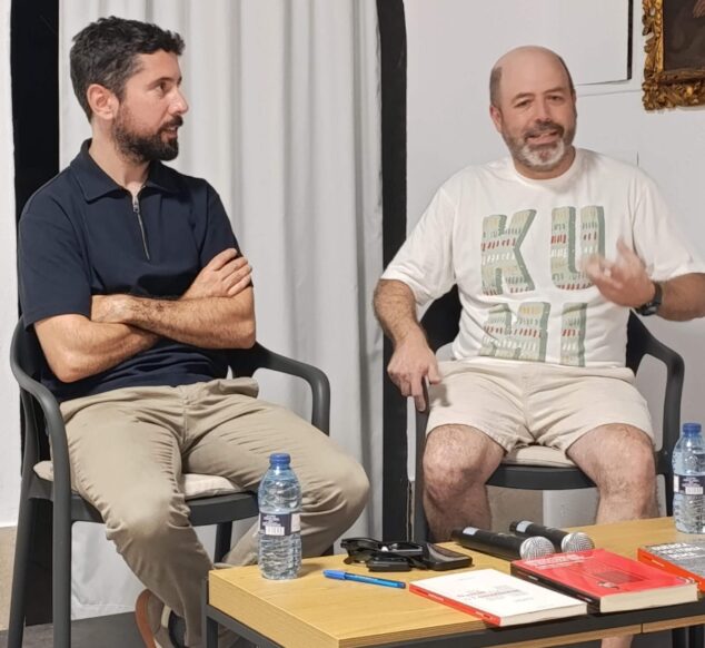 Imagen: Carles Fenollosa y Herme Maria sentados en una mesa con libros y micrófonos durante una presentación literaria.