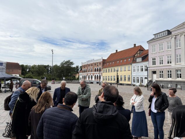 Imagen: Grupo de personas de pie en círculo conversando en una plaza adoquinada rodeada de edificios de arquitectura danesa en Nyborg.