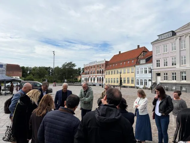 Imagen: Grupo de personas de pie en círculo conversando en una plaza adoquinada rodeada de edificios de arquitectura danesa en Nyborg.
