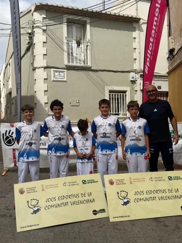 Imagen: Cinco jóvenes jugadores de pilota y su entrenador posan con medallas y diplomas delante de pancartas de los Jocs Esportius de la Comunitat Valenciana.