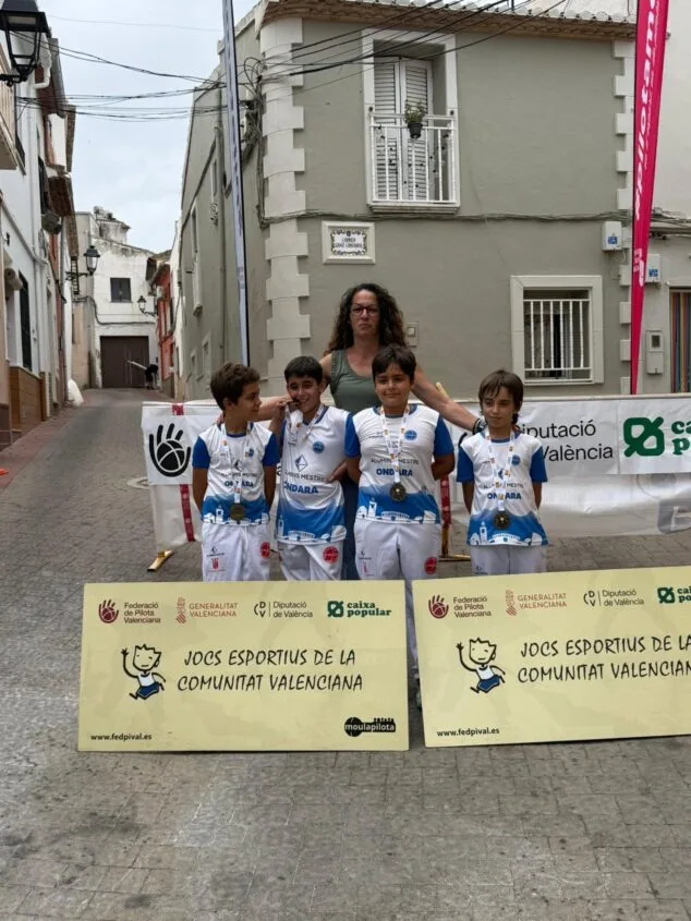Cuatro niños de la Escola de Pilota d'Ondara con uniformes y medallas posan ante carteles de los Jocs Esportius.