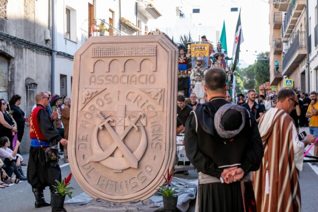Imagen: Escudo de piedra de la Associació de Moros i Cristians de Benissa con festeros detrás.