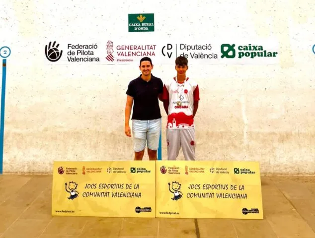 Imagen: Un joven jugador de pilota valenciana de la escuela de Ondara con una medalla y un hombre posan frente a carteles de los Jocs Esportius y patrocinadores.