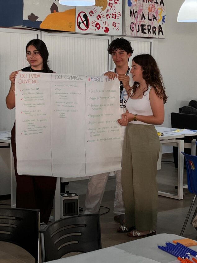 Imagen: Tres jóvenes sostienen un cartel grande con propuestas manuscritas sobre ocio y movilidad durante un foro.