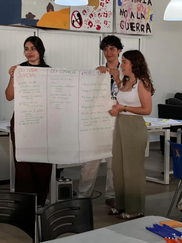 La Xarxa Jove de la Marina Alta fomenta la participación juvenil con un nuevo foro 4 Imagen: Tres jóvenes sostienen un cartel grande con propuestas manuscritas sobre ocio y movilidad durante un foro.