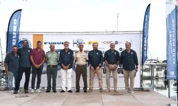 Grupo de siete hombres, algunos uniformados, posando en un escenario exterior delante de un photocall con el logo de "Colosos del Mediterráneo 2026", Club Náutico Moraira, IEO-CSIC y otros patrocinadores.