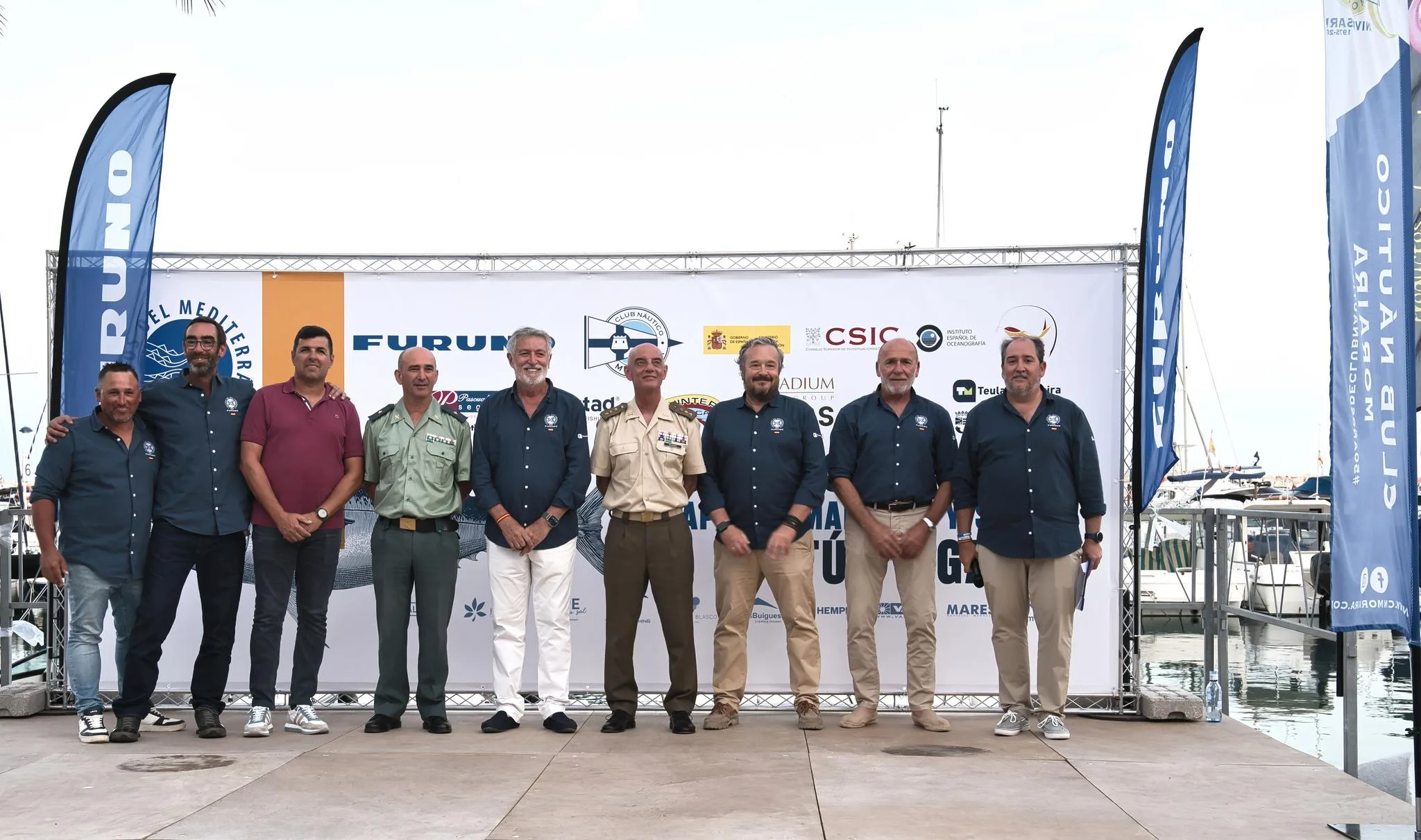 Grupo de siete hombres, algunos uniformados, posando en un escenario exterior delante de un photocall con el logo de "Colosos del Mediterráneo 2026", Club Náutico Moraira, IEO-CSIC y otros patrocinadores.