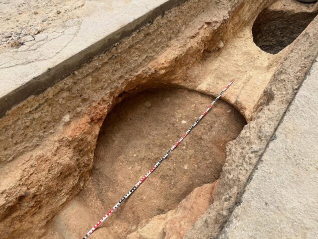 Imagen: Fotografía de una excavación arqueológica en una zanja de obra, mostrando estructuras circulares de tierra y una regla de medición roja y blanca.