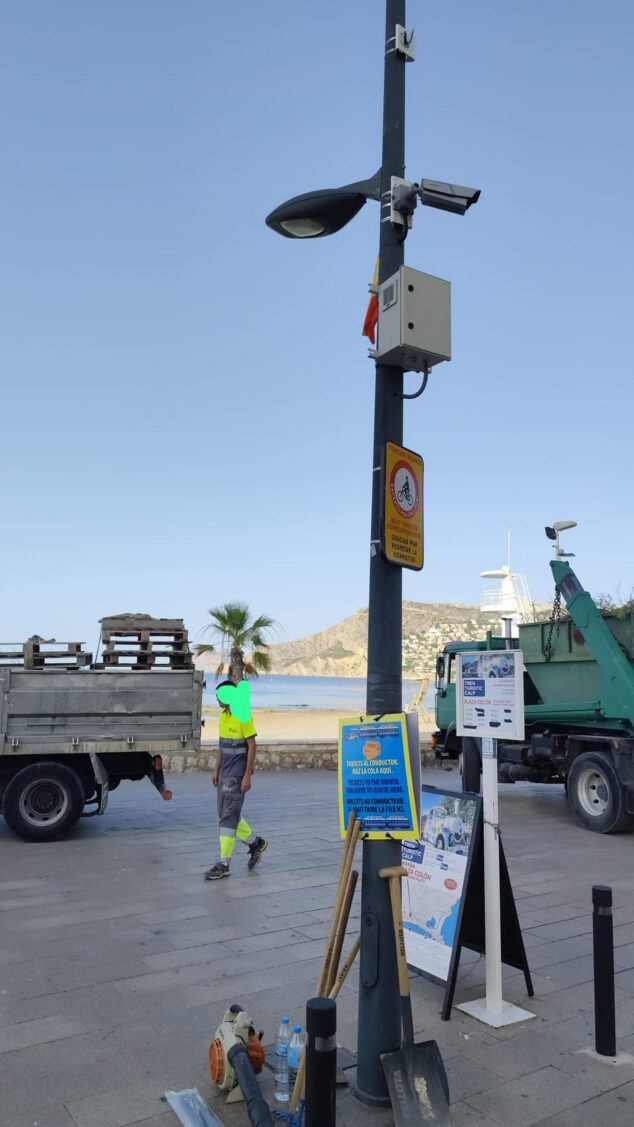 Imagen: Una cámara de videovigilancia montada en un poste cerca de la playa en Calp, con un operario caminando y un camión de obra al fondo.