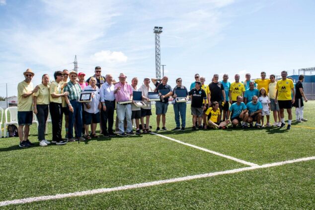 La Jornada Grogueta de la UD Ondarense reúne a aficionados y leyendas en Ondara 2 Imagen: Numeroso grupo de personas posando en un campo de fútbol, algunos sosteniendo placas conmemorativas, durante la Jornada Grogueta de la UD Ondarense.
