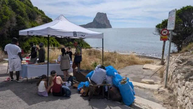 Imagen: Grupo de personas junto a una carpa del Ayuntamiento de Calp y bolsas de basura en una playa con un peñón al fondo.