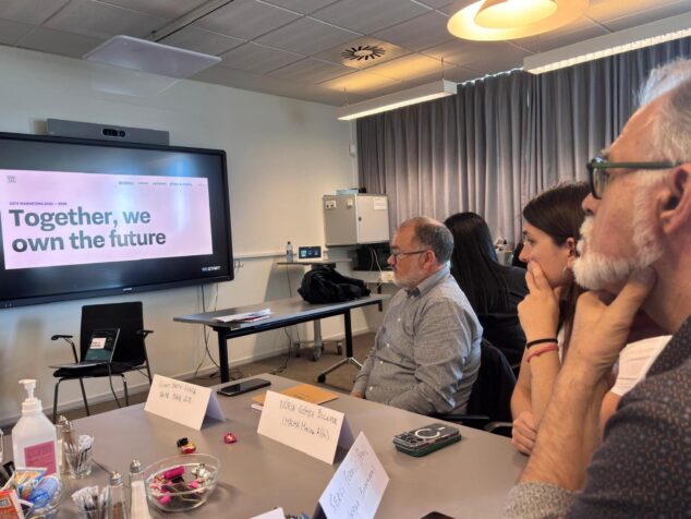 Imagen: Grupo de personas en una sala de reuniones mirando una pantalla con una presentación sobre city marketing durante el encuentro RE:START URBACT.