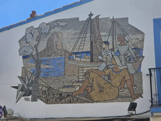 Imagen: Fotografía del gran mosaico mural de Gastón Castelló en una fachada de Calp, que muestra figuras de pescadores y un barco en un paisaje costero.