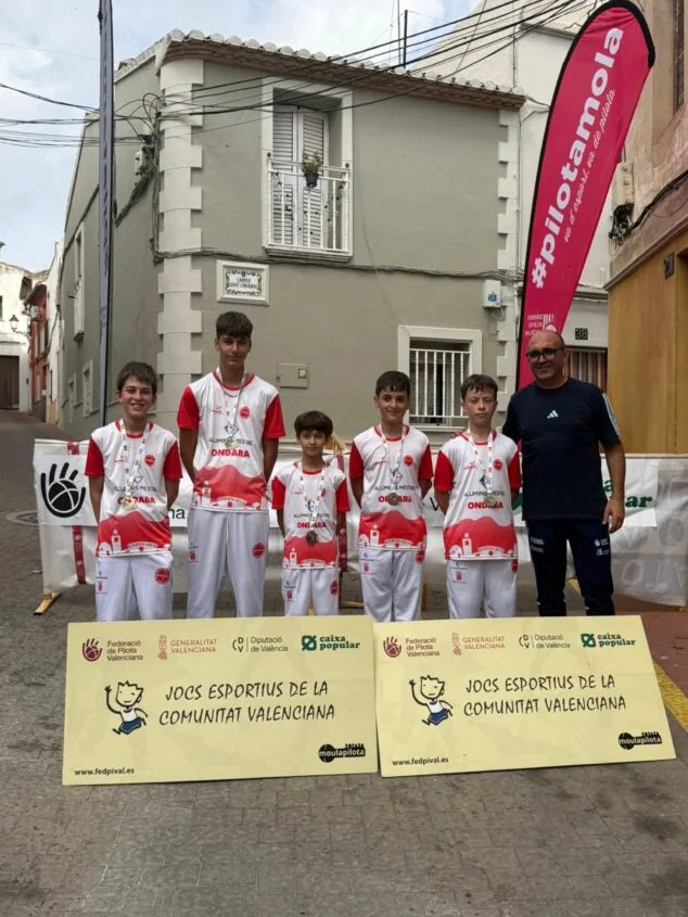 Five boys in Ondara kits with medals and an adult man pose in front of Jocs Esportius de la Comunitat Valenciana banners in a street.