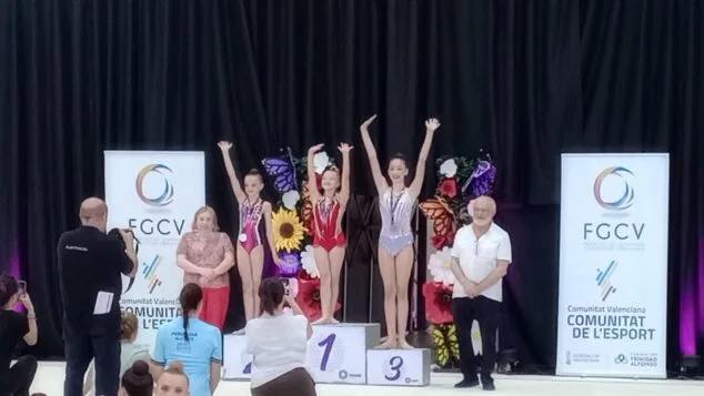 Siete gimnastas del Club Calp competirán en el Campeonato de España 5 Tres gimnastas del Club Calpe en el podio levantando los brazos con medallas, acompañadas por autoridades en una ceremonia de premios de la FGCV.