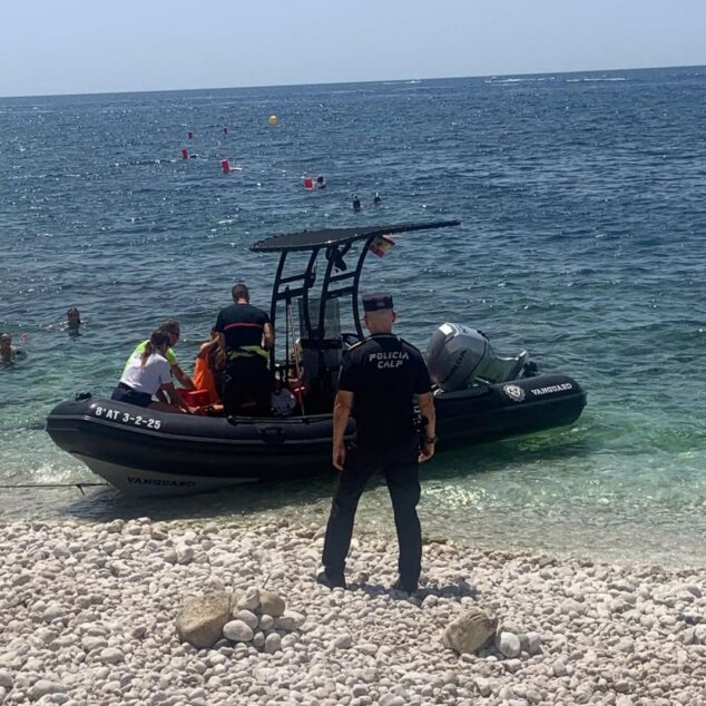 Imagen: Agente de la Policía Local de Calp de espaldas junto a una embarcación semirrígida en la orilla de una playa de piedras.