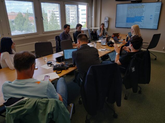 Pedreguer participa en Helsinki en una cumbre europea sobre deporte sostenible 2 Imagen: Varios participantes en una mesa de conferencias durante una reunión de trabajo del proyecto "Olympics of Sustainability" en Helsinki, con una pantalla mostrando información del proyecto.