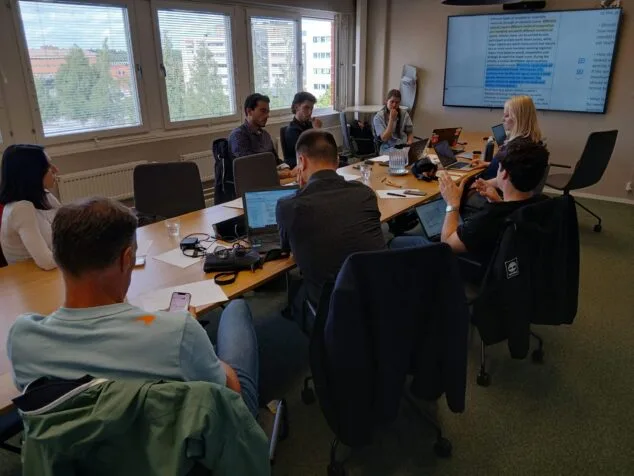 Pedreguer participa en Helsinki en una cumbre europea sobre deporte sostenible 2 Imagen: Varios participantes en una mesa de conferencias durante una reunión de trabajo del proyecto "Olympics of Sustainability" en Helsinki, con una pantalla mostrando información del proyecto.