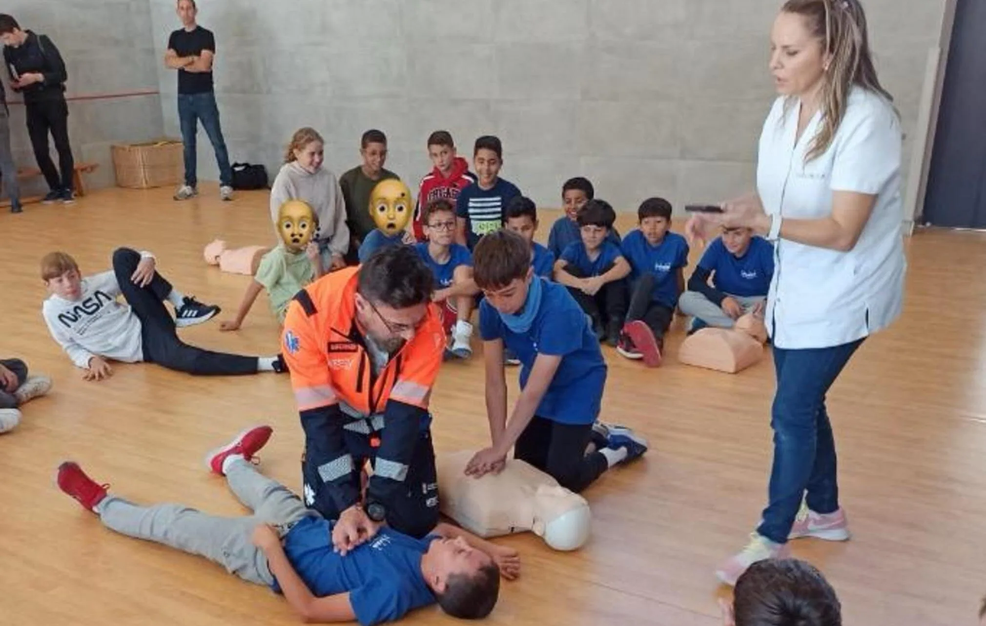 Un profesional sanitario enseña a un alumno a hacer RCP en el suelo de un gimnasio, mientras otros niños y una enfermera observan.