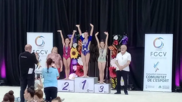 Siete gimnastas del Club Calp competirán en el Campeonato de España 2 Imagen: Tres gimnastas jóvenes, incluyendo a Zoe Noriega en el centro, están de pie en un podio con medallas y los brazos levantados, flanqueadas por dos autoridades en un evento de la FGCV.