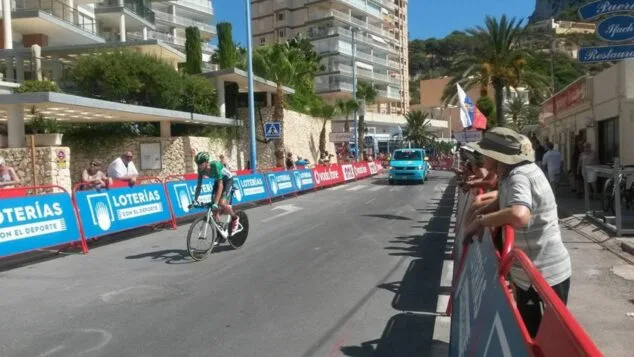 Un ciclista profesional sobre una bicicleta de contra reloj pedalea por una calle urbana de Calp delimitada con vallas de patrocinadores de La Vuelta y con público observando.