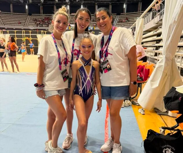 Imagen: Una joven gimnasta con maillot posa junto a tres mujeres adultas con camisetas del Club Les Marines en el interior de un pabellón deportivo.