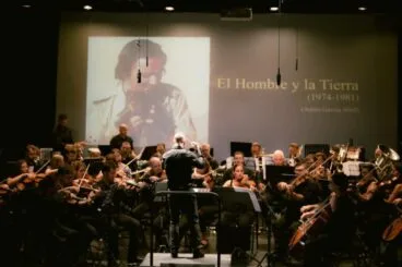 Orquesta sinfónica tocando en un escenario con una gran pantalla de fondo que muestra una imagen de Antón García Abril y el texto "El Hombre y la Tierra".