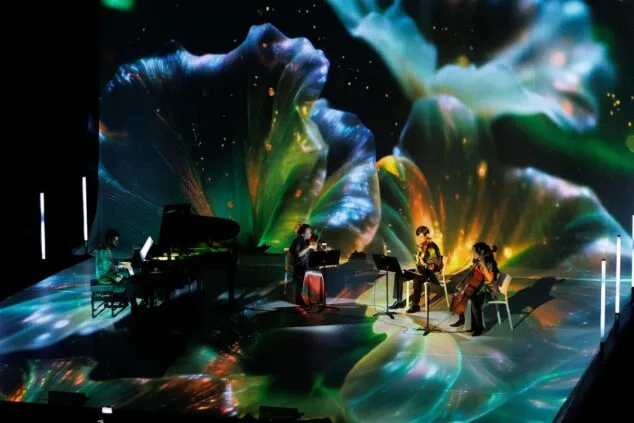 Músicos tocando piano, violín y violonchelo en un escenario envuelto por proyecciones visuales coloridas durante el concierto inmersivo Colors del So en Calp.