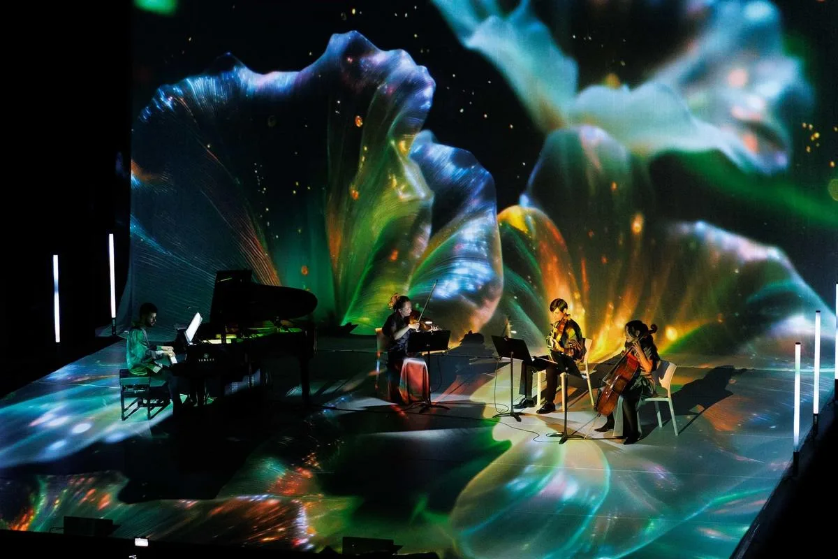 Músicos tocando piano, violín y violonchelo en un escenario envuelto por proyecciones visuales coloridas durante el concierto inmersivo Colors del So en Calp.