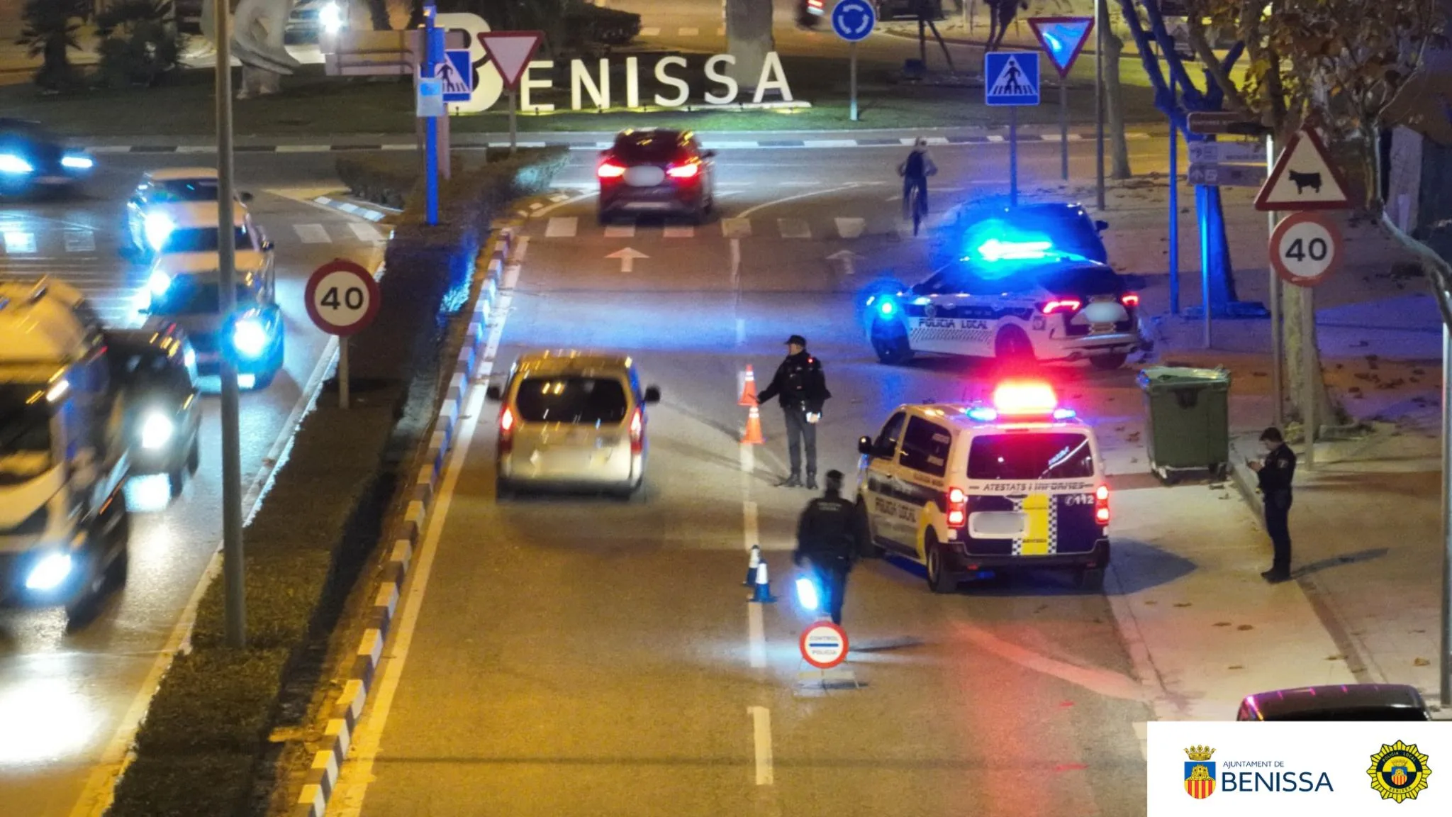 Agentes de la Policía Local de Benissa en un control nocturno con coches patrulla y luces azules encendidas cerca de una rotonda con el nombre del municipio.