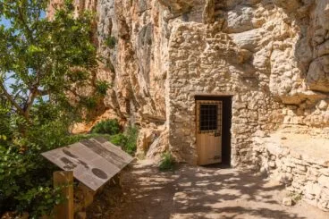 Entrada de piedra y puerta de madera de la Cova de les Morretes con un panel informativo al lado.