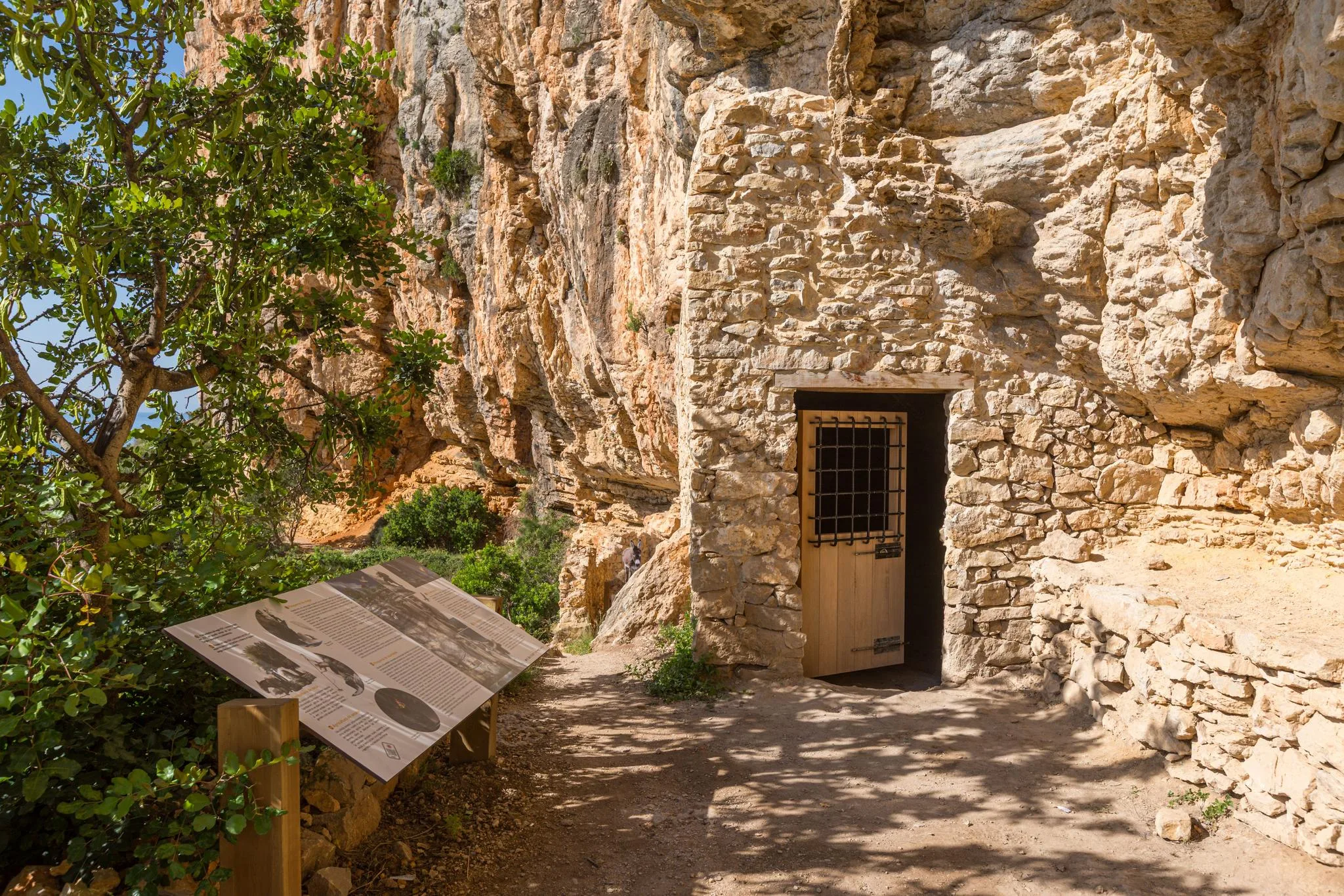 Entrada de piedra y puerta de madera de la Cova de les Morretes con un panel informativo al lado.
