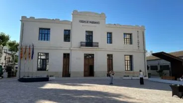 Fotografía exterior de la fachada de piedra del edificio de las Escuelas Graduadas y Ajuntament de Benissa, con banderas oficiales en la entrada.