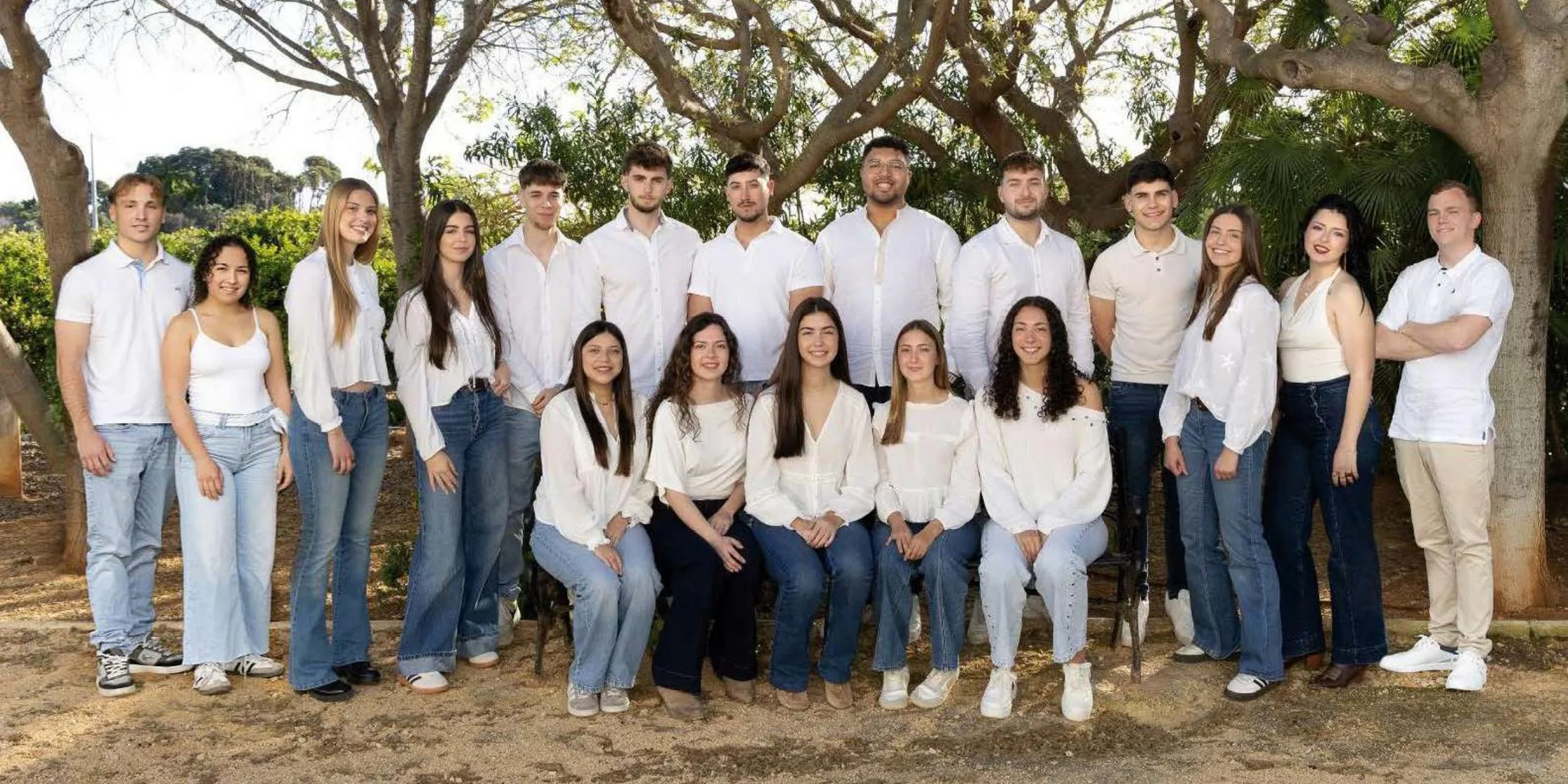 Foto de grupo de los jóvenes Festeros 2026 de Benitatxell, vestidos de blanco y vaqueros en un entorno exterior con árboles.