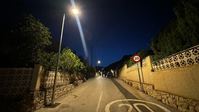 Más luz en Calp: 67 nuevas farolas solares iluminan sus urbanizaciones 4 Imagen: Vista nocturna de una calle de urbanización en Calp iluminada por una farola solar, con dos figuras caminando en la distancia.