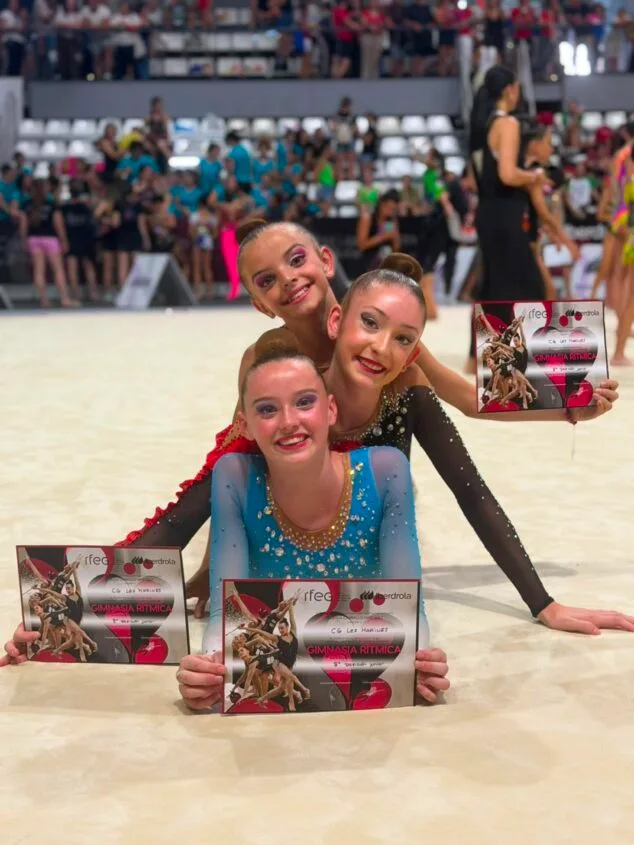 Tres gimnastas del Club Les Marines sostienen diplomas en un tapiz de competición.